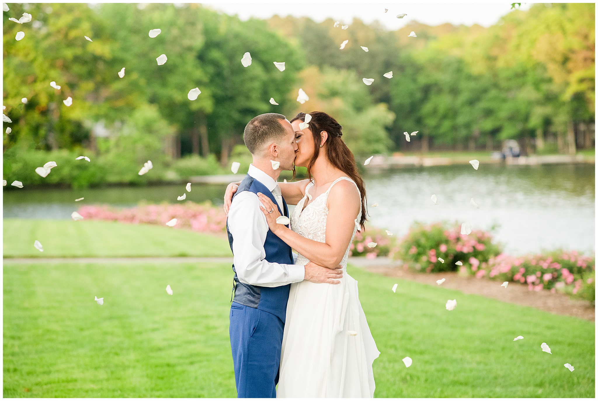 Newlyweds kiss in a manicured lawn by a lake while white flower petals rain down around them Wedding Venues in Virginia