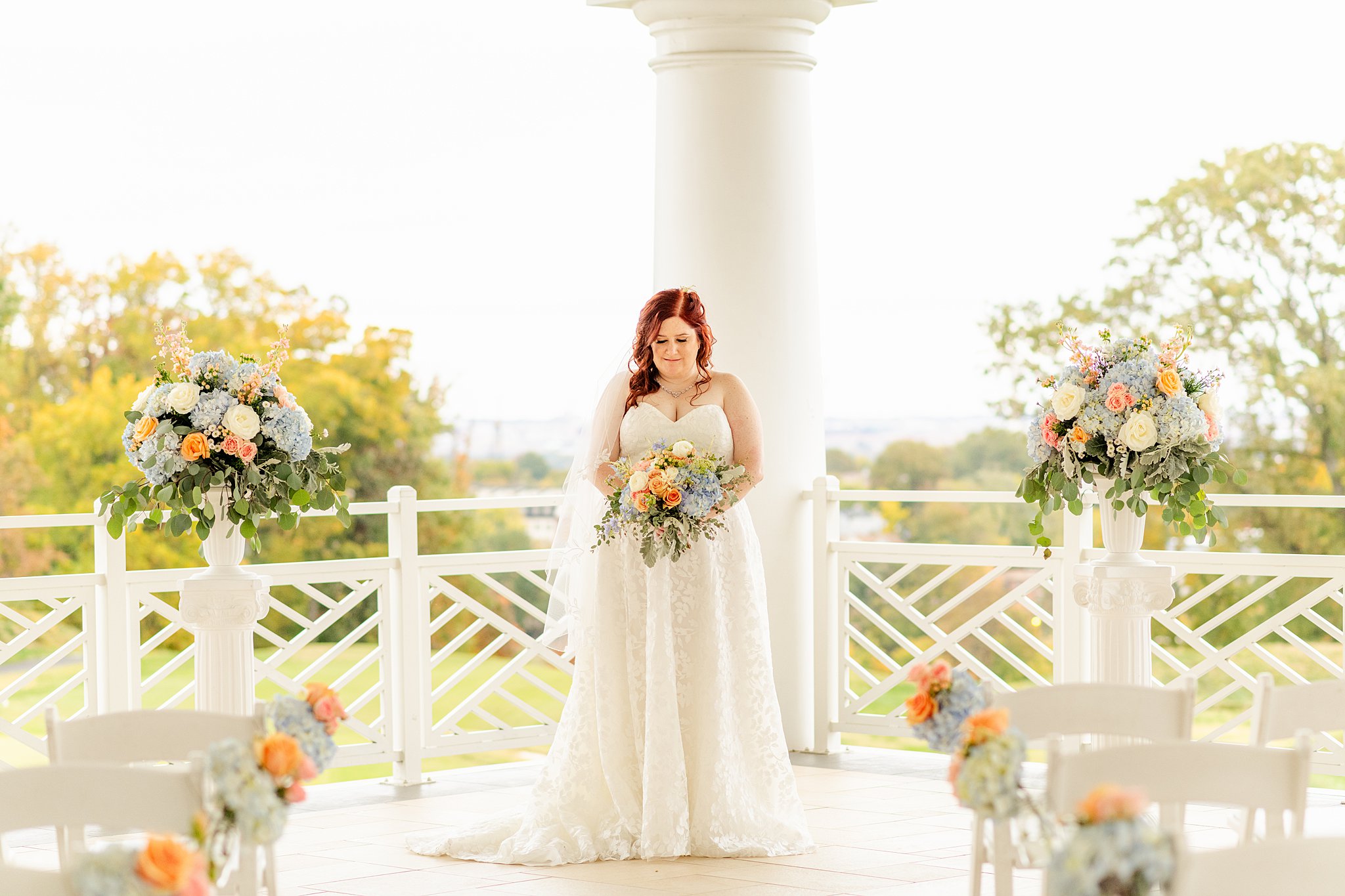 A bride in a long white lace dress stands at the front of her ceremony location with matching bouquet and floral decorations in front of a large column wedding venues in northern virginia