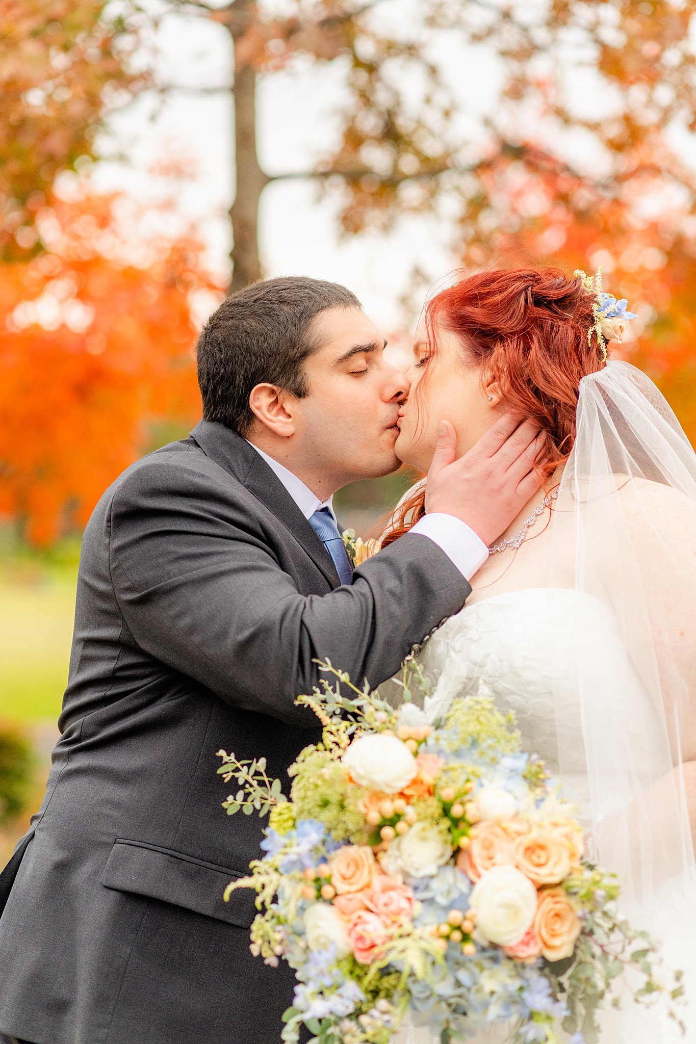 Newlyweds kiss in a park with fall colored trees and a large bouquet