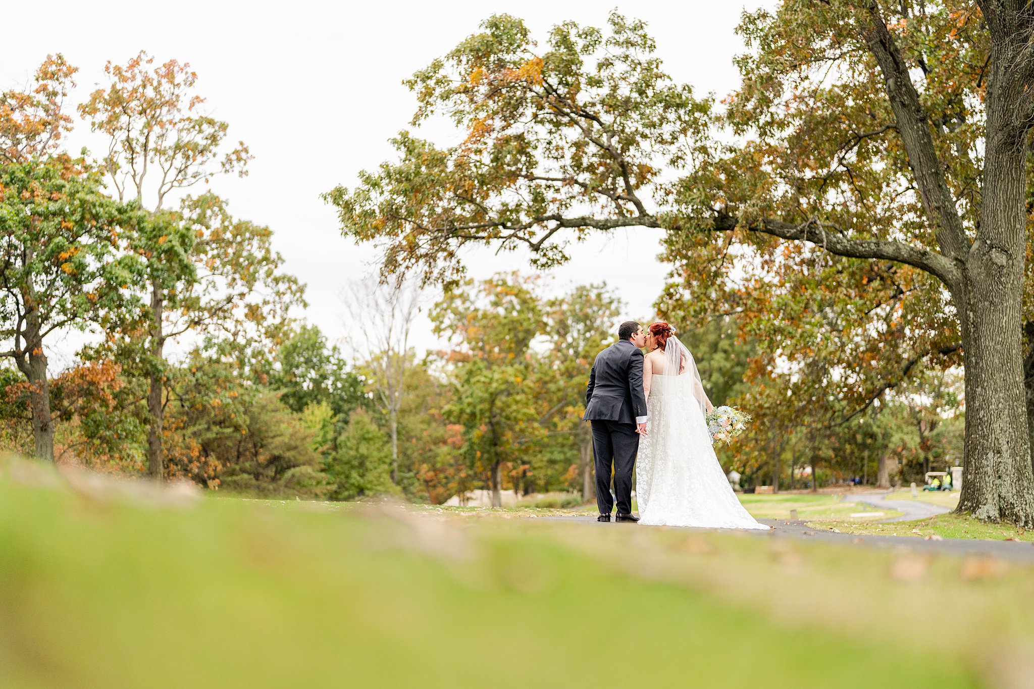 Newlyweds kiss under a tall tree while walking down a park path