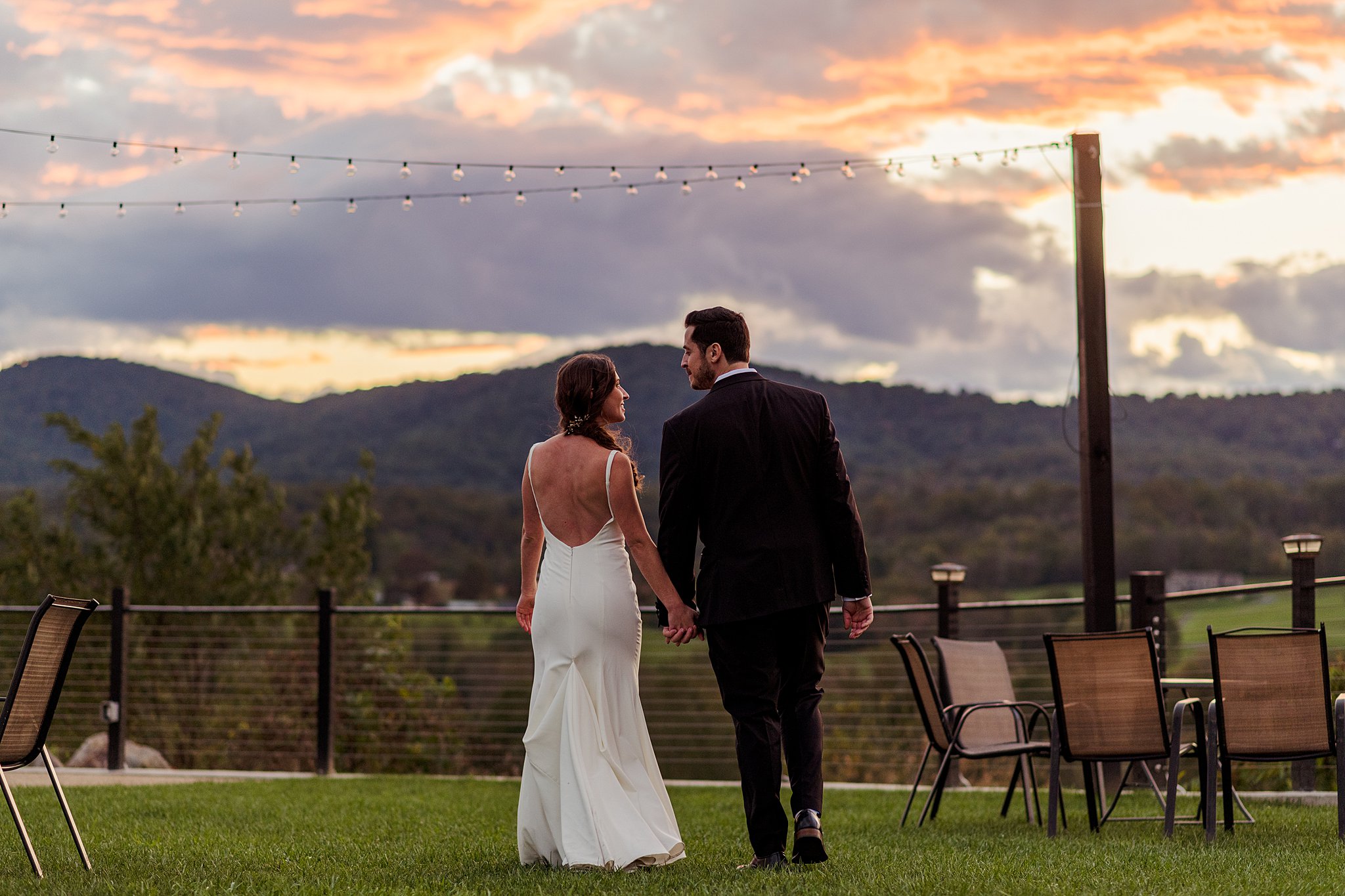 Newlyweds hold hands while looking at each other on a lawn with chairs and a mountain sunset in the background wedding venues in northern virginia