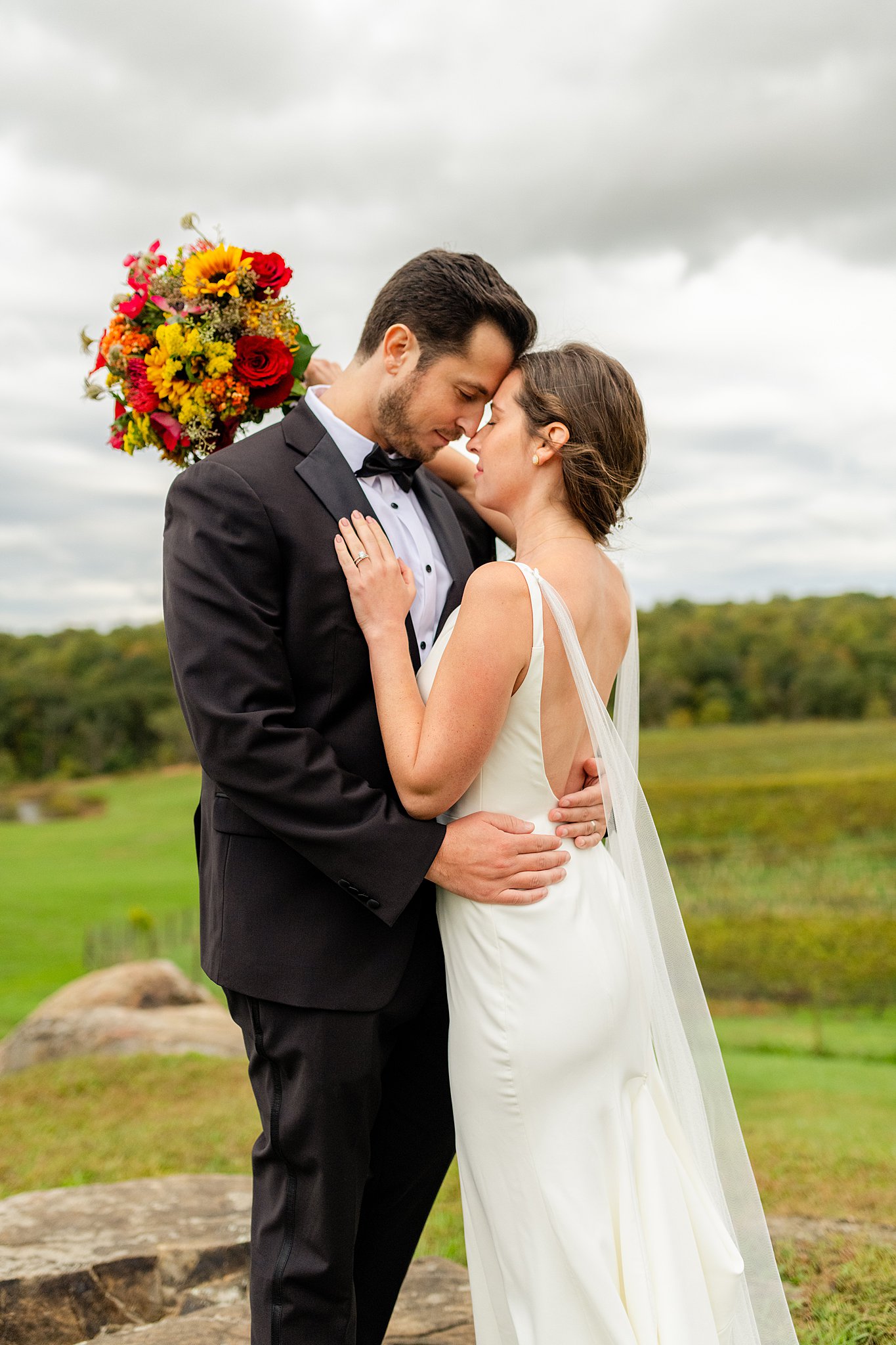 Newlyweds hug nose to nose on a rocky hill above a vineyard with a colorful bouquet wedding venues in northern virginia